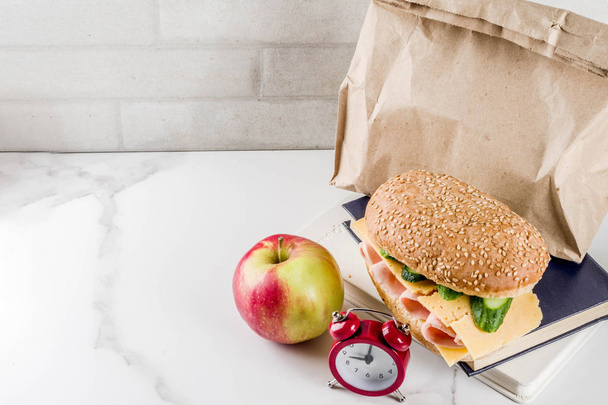 Zdrowa Szkoła jedzenie koncepcja, papierowa torba z lunchu, apple, sandwich, książki i budzik na przestrzeni kopii tabeli biała kuchnia - Zdjęcie, obraz