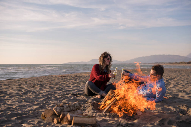Young Couple Relaxing By The Fire, Drinking A Beer Or A Drink From The Bottle on the beach at autumn day - Photo, image