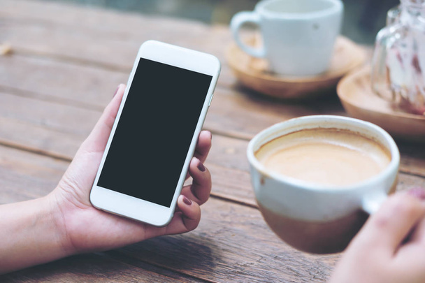 Mockup image of white mobile phone with blank black screen and hot coffee on vintage wood table in cafe - Photo, Image