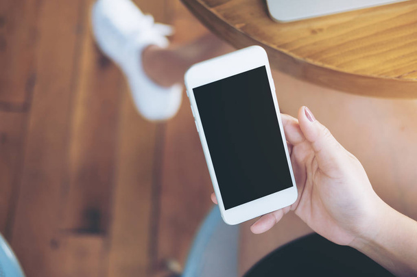 Imagen burlona de manos sosteniendo teléfono móvil blanco con pantalla negra en blanco en el muslo con portátil de color plata en la mesa y fondo de madera
 - Foto, Imagen