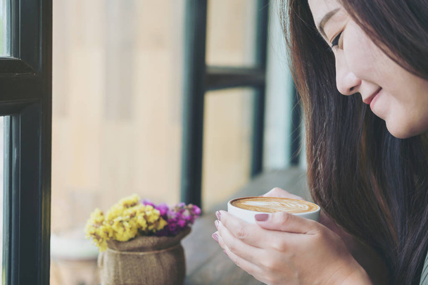 Μια όμορφη Ασιάτισσα κρατώντας και μυρίζοντας ζεστό φλιτζάνι καφέ σε loft cafe με αίσθημα ευτυχίας και χαμογελαστό πρόσωπο - Φωτογραφία, εικόνα
