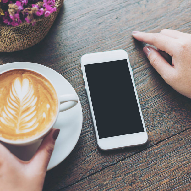 Mockup image of white mobile phone with blank black screen and hand holding hot latte coffee on vintage wood table in cafe - Photo, Image