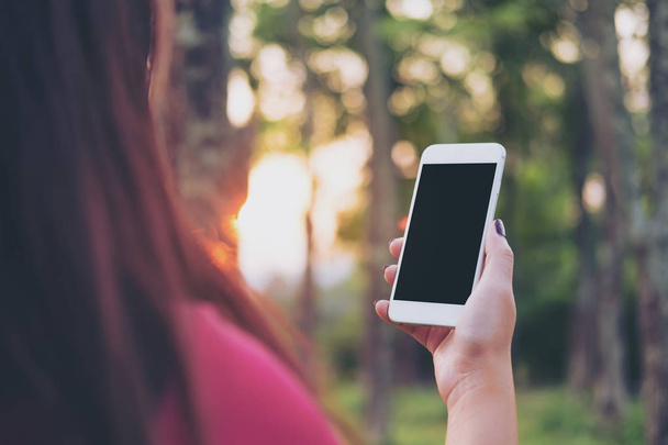 Mockup beeld van een vrouw te houden en met behulp van slimme telefoon met leeg zwart scherm bij outdoor en groene natuur achtergrond tijdens zonsondergang tijd - Foto, afbeelding