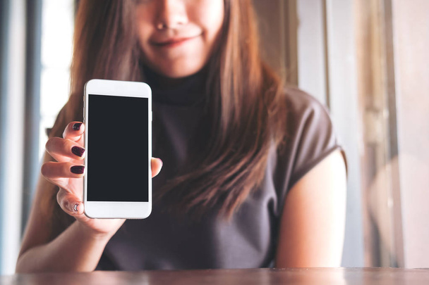 Κοροϊδεύω εικόνα του μια όμορφη γυναίκα με χαμογελαστό πρόσωπο κρατώντας και παρουσιάζουν λευκό κινητό τηλέφωνο με κενή μαύρη οθόνη στο vintage ξύλινο καφέ - Φωτογραφία, εικόνα