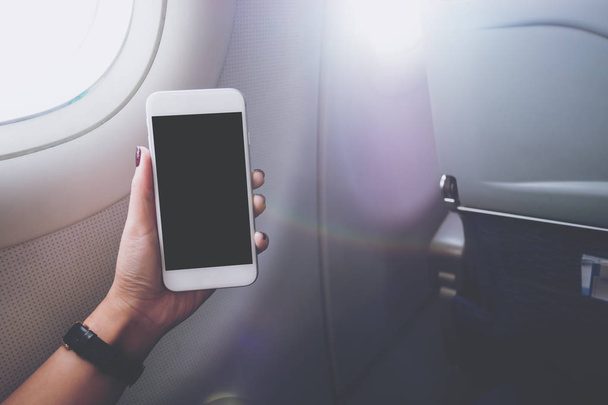 Imagen burlona de una mano sosteniendo y mirando un teléfono inteligente blanco con pantalla negra junto a una ventana de avión con nubes y fondo de cielo
 - Foto, Imagen