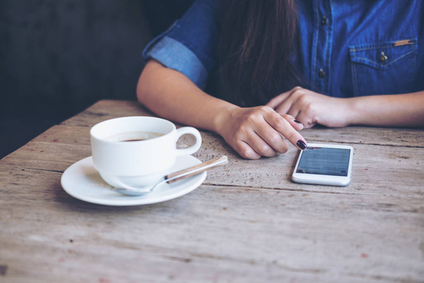女性の感覚でスマート フォンを使用して木製のテーブルの上にコーヒー カップを持つヴィンテージのカフェに座りながらリラックス - 写真・画像