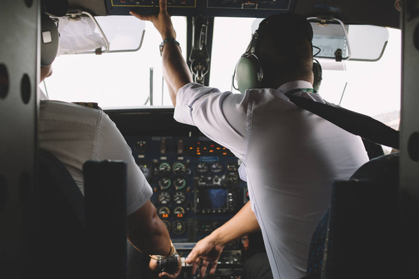 Rear view of two professional pilots dressed in white uniform sitting in private cabin of the plane to control cockpit engine of airplane in air. Flight desk control panel during take off or landing - Photo, Image