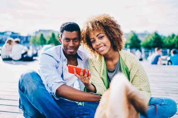 Portrait of smiling afro american couple in love installing new application on smartphone dark skinned friends booking tickets on online on mobile looking at camera resting with dog in city square - Фото, зображення