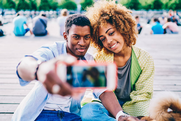 Selective focus on young afro american friends posing while talking picture on modern smartphone camera sitting with dog in park, dark-skinned couple using mobile for making photo resting together - Фото, изображение