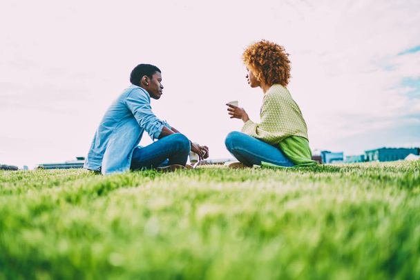 Pensive male and female dark skinned friends sitting on grass talking enjoying break outdoors, trendy dressed afro american hipsters having conversation during free time resting together on meeting - Photo, Image