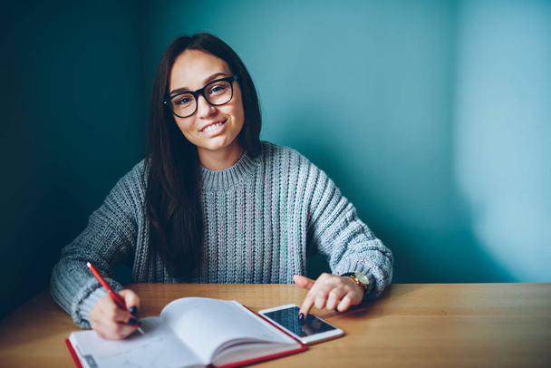Half length portrait of smart cheerful student in eyewear writing planning daily schedule and making to do list in notepad while received notification on smartphone.Publicity area for your advertising text - Фото, зображення