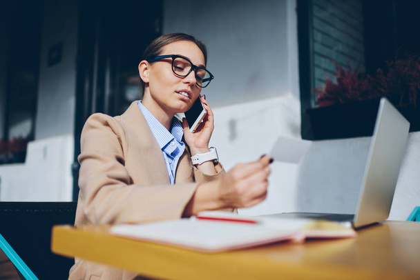 Skilled young woman in spectacles calling phone number from business card sitting at laptop computer connected to wireless internet in searching new job.Smart female talking on smartphone about work - 写真・画像