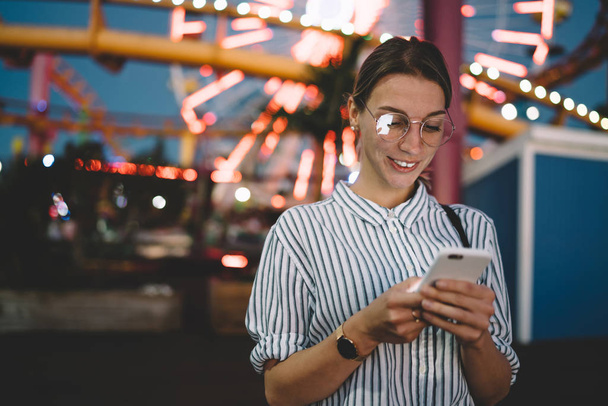 Happy young woman watching funny video online on telephone visiting amusement park with night lights at evening, cheerful hipster girl reading content in networks on cellular resting at evening - Фото, зображення