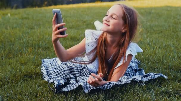 Young beautiful girl making selfie portrait using smartphone in green park, lying on the grass with mobile phone. Color graded - Materiaali, video