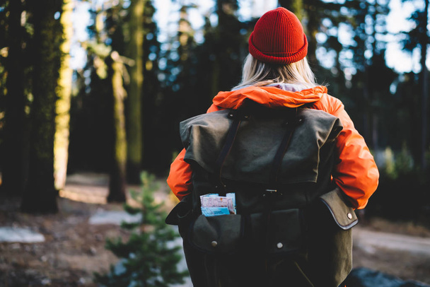 Back view of female traveler in colourful orange jacket recreating during hiking tour in forest, girl wanderlust with touristic rucksack and backpack explore wild environment of wood on active res - Foto, imagen
