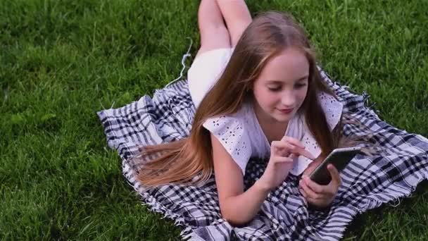 Happy girl typing sms by phone lies on grass in the park - Materiaali, video