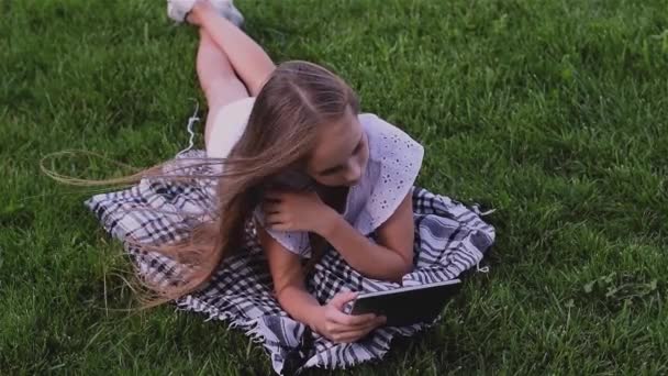 Happy girl using tablet pc lies on grass in the park - Materiał filmowy, wideo