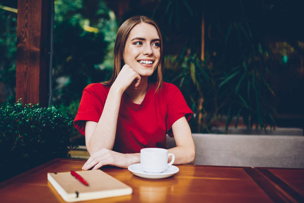 Positive attractive hipster girl dressed in stylish casual outfit looking away and smiling during leisure time in cafe outdoors.Cheerful young entrepreneur resting in coffee shop with cup of cappuccino - Foto, imagen
