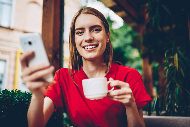 Half length portrait of positive blogger updating multimedia and sharing posts on own website via high speed internet connection on mobile phone while drinking coffee and smiling at camera - 写真・画像