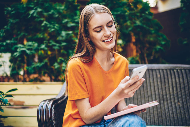 Smiling young woman making payment online via free high speed inernet connection on smartphone device sitting with favourite book outdoors.Cheerful female blogger updating profile on cellular - Photo, image