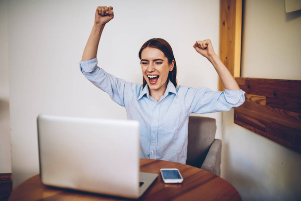 Carefree hipster girl raising hands and screaming celebrating winning in online contest on website sitting at laptop computer connecte to wireless internet sitting in stylish coworking space - Foto, Imagen