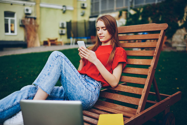 Casual dressed hipster girl resting outdoors on wooden chair and messaging in online chat with friends and followers via free 4g internet connection on smartphone after remotely work at laptop device - Foto, Imagen