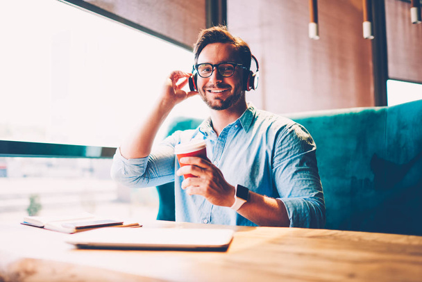 Half length portrait of positive businessman in spectacles listening electronic music in digital headphones enjoying tasty coffee beverage sitting at table in coffee shop interior i recreation time - Fotografie, Obrázek