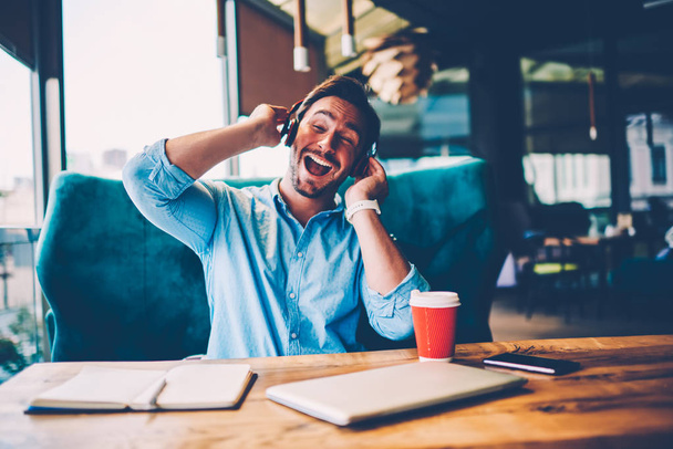 Happy businessman singing favourite song head in modern headphones sitting at table with coffee and notepad in cafe.Positive owner of coffee shop enjoying stereo music during recreation time - Foto, Imagem