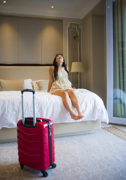 lifestyle portrait of young happy and beautiful Asian Korean tourist woman arriving to luxury hotel room playful on bed smiling cheerful staring around delighted and excited with suitcase - 写真・画像