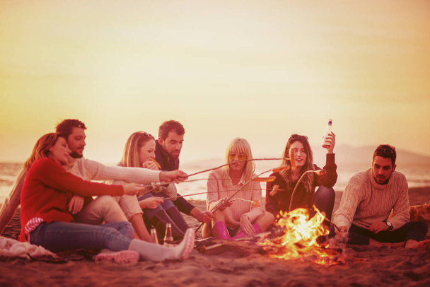 Group of young friends sitting by the fire at autumn beach, grilling sausages and drinking beer, talking and having fun filter - Фото, зображення