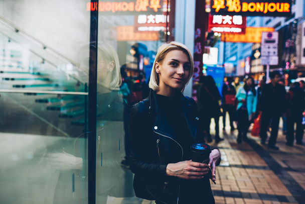 Cute dreamy young woman with short haircut walking on street and enjoying trip.Beautiful blonde hipster girl dressed in leather jacket holding coffee to go in hands while strolling in urban setting - Photo, image