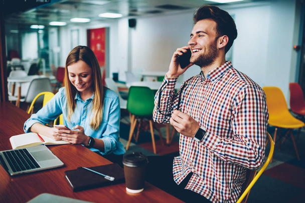 Bearded man laughing during mobile conversation with friend on smartphone while sitting in coworking with hipster girl.Young man chatting online on telephone while boyfriend talking on cellular - Foto, Imagem