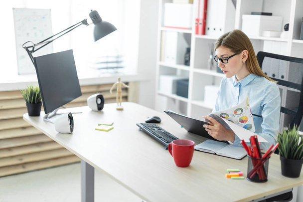 Une jeune fille à lunettes s'assoit à une table dans le bureau, tient un crayon à la main et travaille avec des documents
. - Photo, image