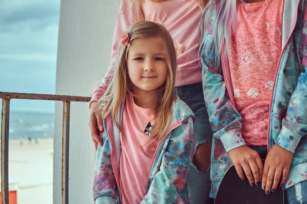 Portrait d'une jolie petite fille aux cheveux blonds vêtue d'un t-shirt rose et d'un sweat à capuche bleu debout avec sa famille près d'un garde-corps contre la côte de la mer
. - Photo, image