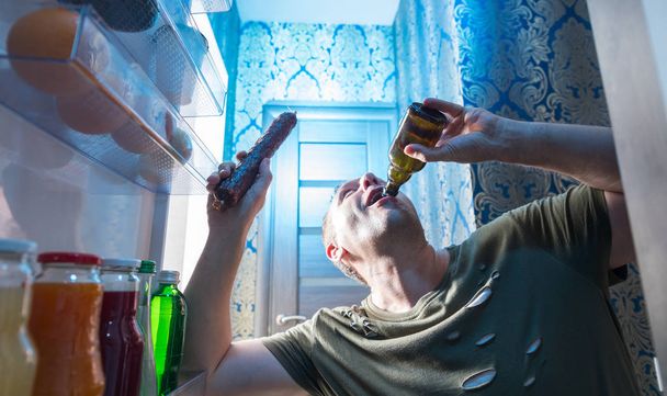 Man slugging back a bottle of beer from his fridge while holding a whole salami in his other hand viewed from inside the refrigerator - Foto, imagen