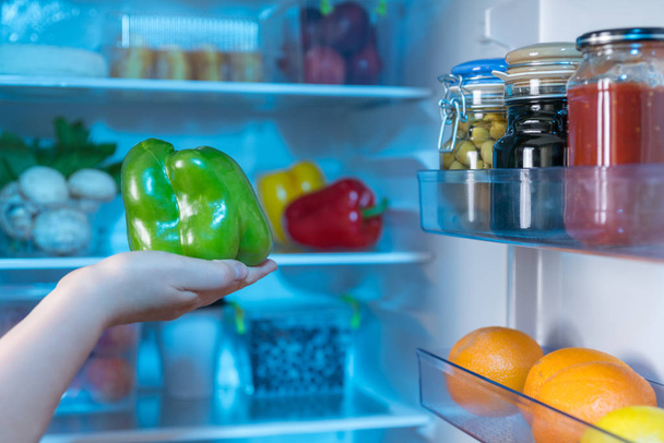 Person holding fresh green pepper against open refrigerator - Fotó, kép