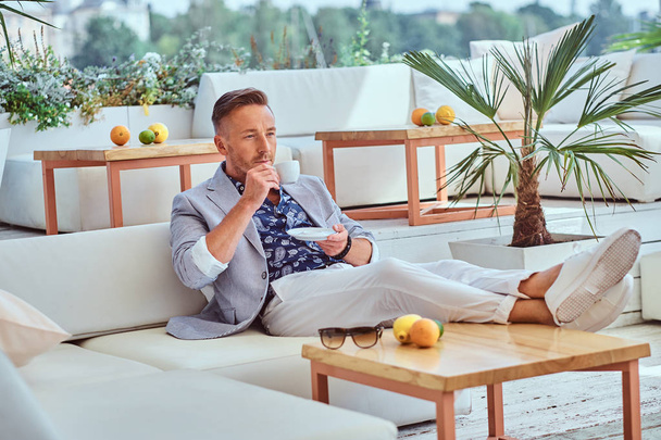 Fashionable successful man with stylish hair dressed in modern elegant clothes holds a cup of coffee while sitting on a sofa at outdoor cafe against the background of the city wharf. - 写真・画像