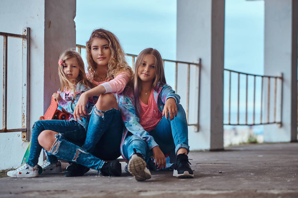 Beautiful girl with blonde hair and her little sisters dressed in trendy clothes sitting together on skateboards posing near a guardrail against a sea coast. - Fotó, kép