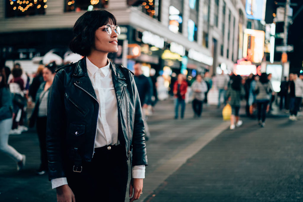 Stylish hipster girl dressed in trendy jacket walking outdoors on avenue of metropolis with crowd of people.Brunette young woman in eyeglasses looking away standing on New York street et evening - Zdjęcie, obraz