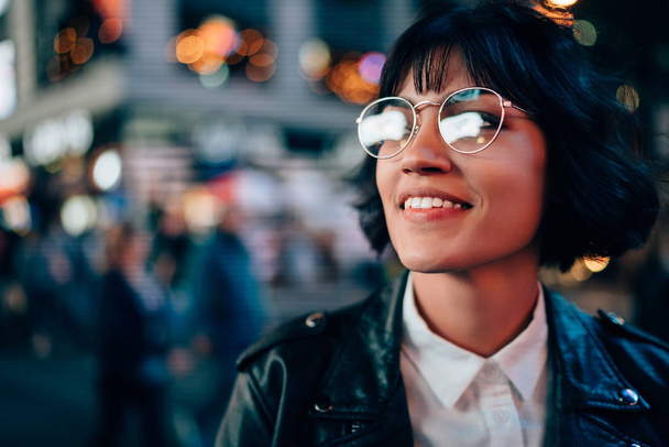 Charming positive brunette young woman in cool spectacles enjoying night street standing on bokeh background.Cheerful attractive hipster girl in stylish eyeglasses and leather jacket laughing - Fotoğraf, Görsel