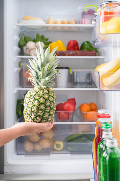 Person holding fresh pineapple against open fridge full of food - Foto, afbeelding