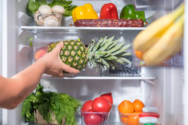 Man putting fresh pineapple into refrigerator full of food - Valokuva, kuva