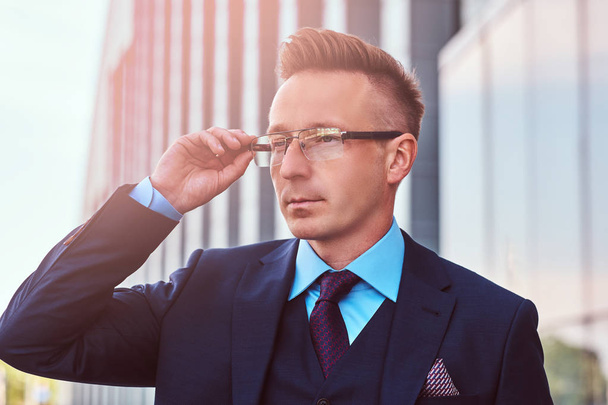 Close-up portret van een vertrouwen stijlvolle zakenman gekleed in een elegante pak weg op zoek en zijn bril kunnen corrigeren terwijl permanent buiten tegen de achtergrond van een wolkenkrabber. - Foto, afbeelding