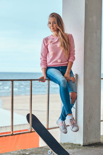 Affascinante donna con i capelli biondi vestita con abiti alla moda seduta su un guardrail contro una costa di mare
. - Foto, immagini