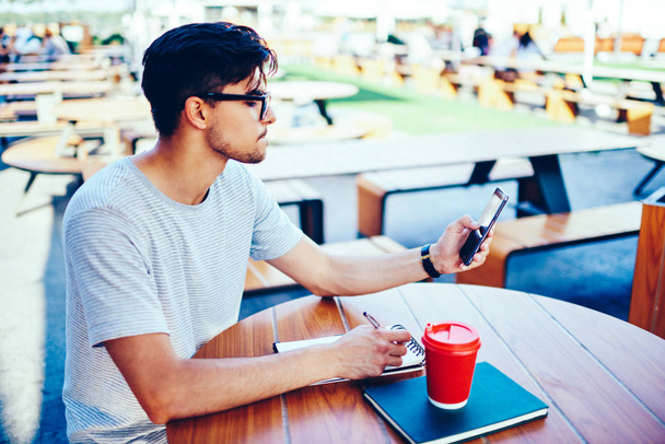 Düşünceli erkek blogger rahat t-hirt ve şık gözlük ders kitabı ve kahve fincanı ile tahta masada oturan smartphone ücretsiz 4 g internet bağlantısı kullanarak Web sitesinden metin bilgileri yeniden yazma - Fotoğraf, Görsel