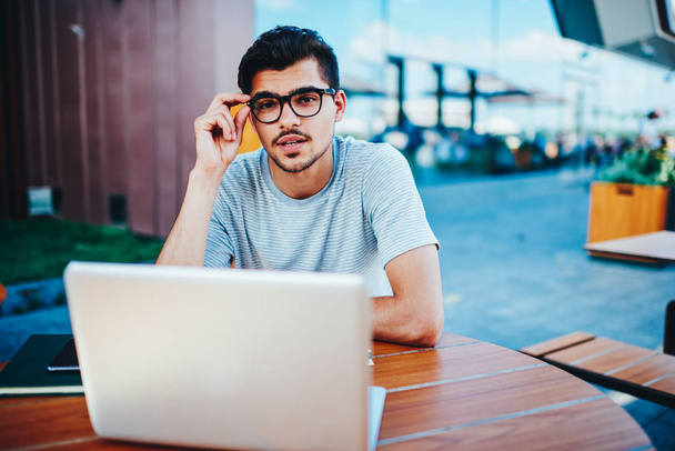 Half length portrait of stylish young student in optical eyeglasses looking at camera while sitting at modern laptop device outdoors.Handsome hipster guy in spectacles with netbook - Photo, Image
