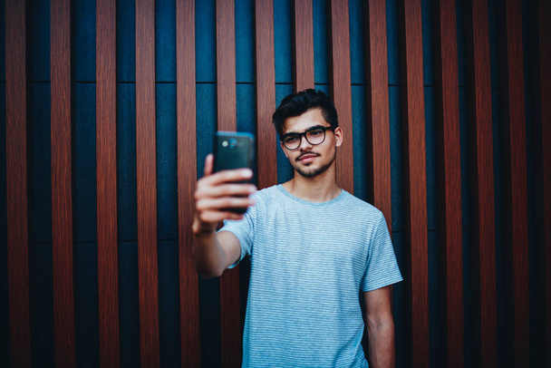 Male blogger in stylish eyeglasses shotting video of front camera of modern smartphone for followers standing outdoors promotional background.Hipster guy in t-shirt making selfie photos on telephone - Fotografie, Obrázek