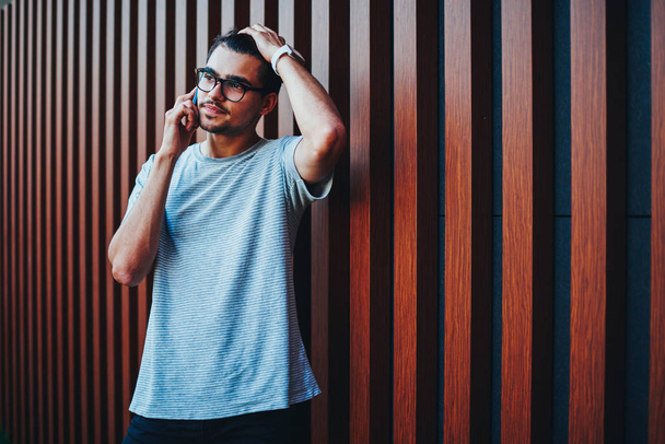 Casual dressed hipster bloger in eyeglasses having mobile conversation on smartphone device standing outdoors near wooden wall.Young man in trendy t-shirt talking with operator on cellular - Valokuva, kuva