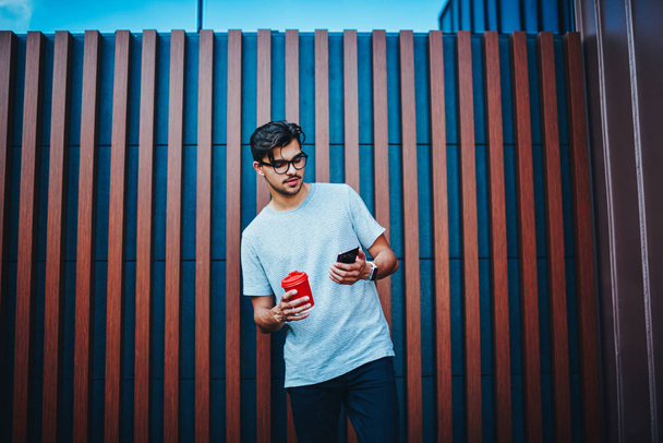 Stylish male blogger checking mail in networks on smartphone connected to 4G internet standing with coffee to go in urban setting.Hipster guy in eyewear chatting online on promotional background - Fotoğraf, Görsel