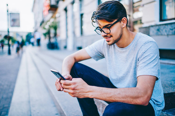 Cheerful male bogger reading funny comments from followers on website on smartphone with 4G internet.Positive hipster guy in eyewear reading notification with good new on telephone sitting in downtown - Фото, зображення